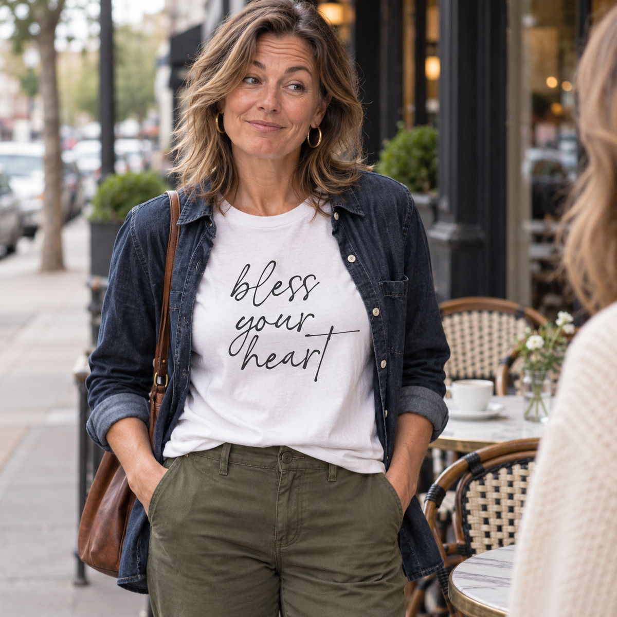 Woman in a white Bless Your Heart tee and denim overshirt outside a cafe.