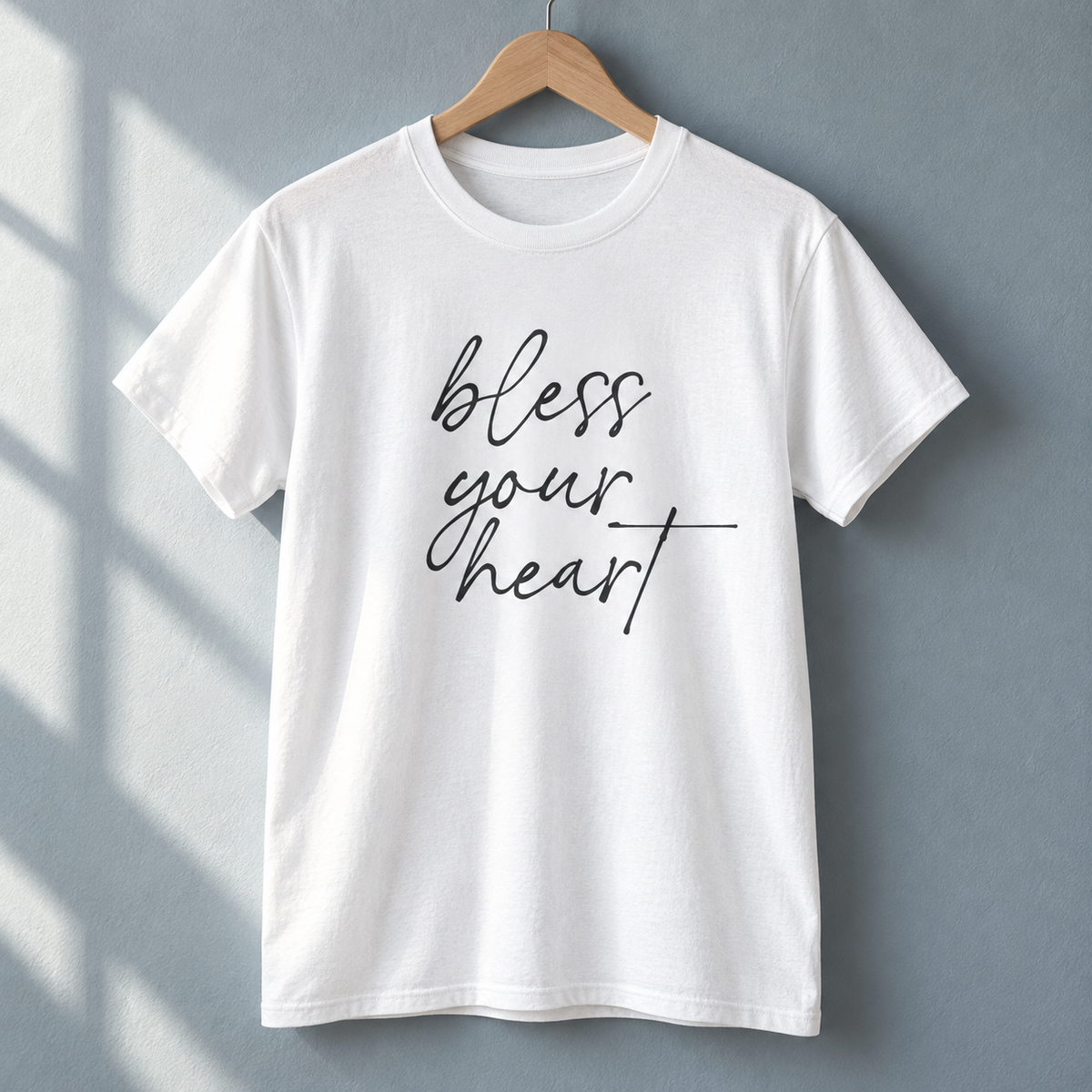 White Bless Your Heart script tee hanging against a blue-gray wall.