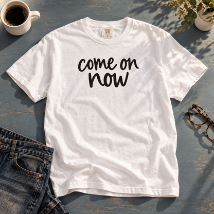 White Come On Now tee laid with jeans, coffee, and glasses on a blue-gray table.
