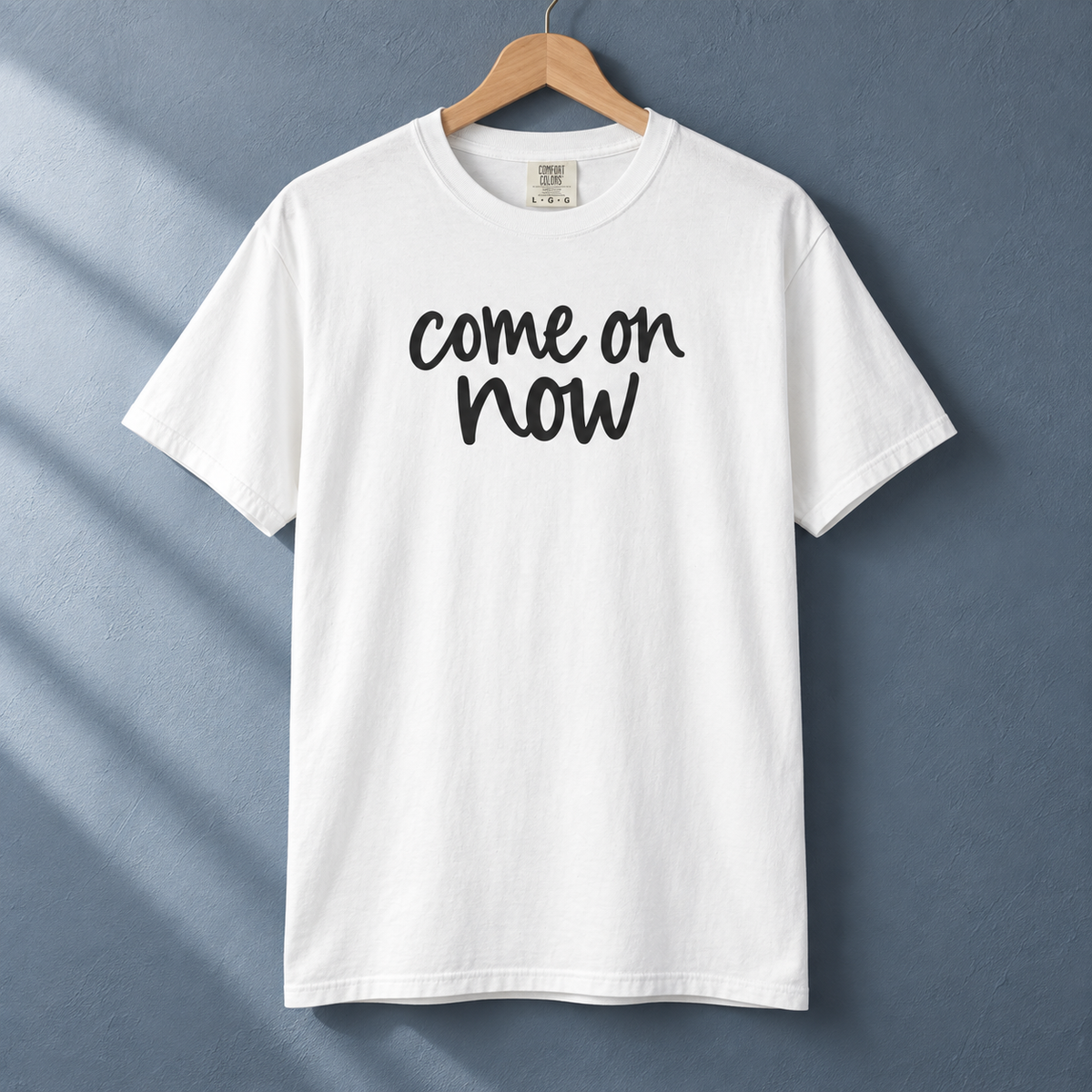 White Come On Now graphic tee hanging against a slate blue wall.