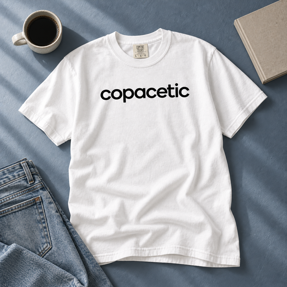 White Copacetic tee laid beside jeans, coffee, and a book on a blue-gray table.