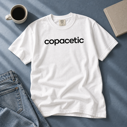 White Copacetic tee laid beside jeans, coffee, and a book on a blue-gray table.