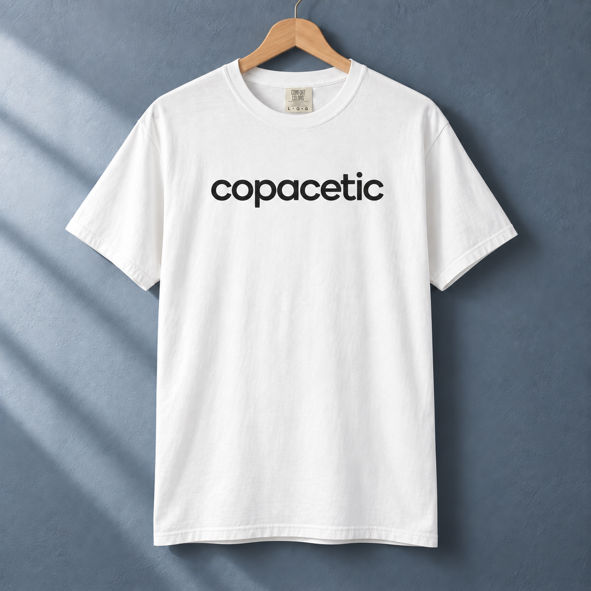 White Copacetic graphic tee on a wood hanger against a blue-gray wall.