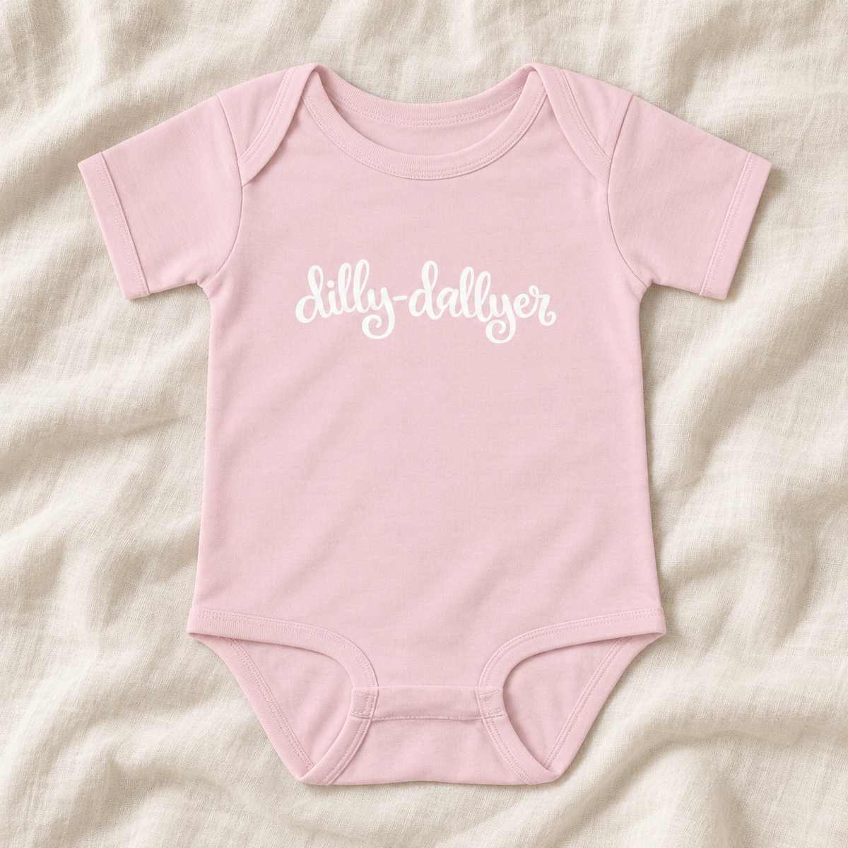 Ballerina baby bodysuit with white “dilly-dallyer” script, laid flat on ivory linen