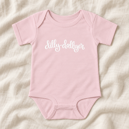 Ballerina baby bodysuit with white “dilly-dallyer” script, laid flat on ivory linen