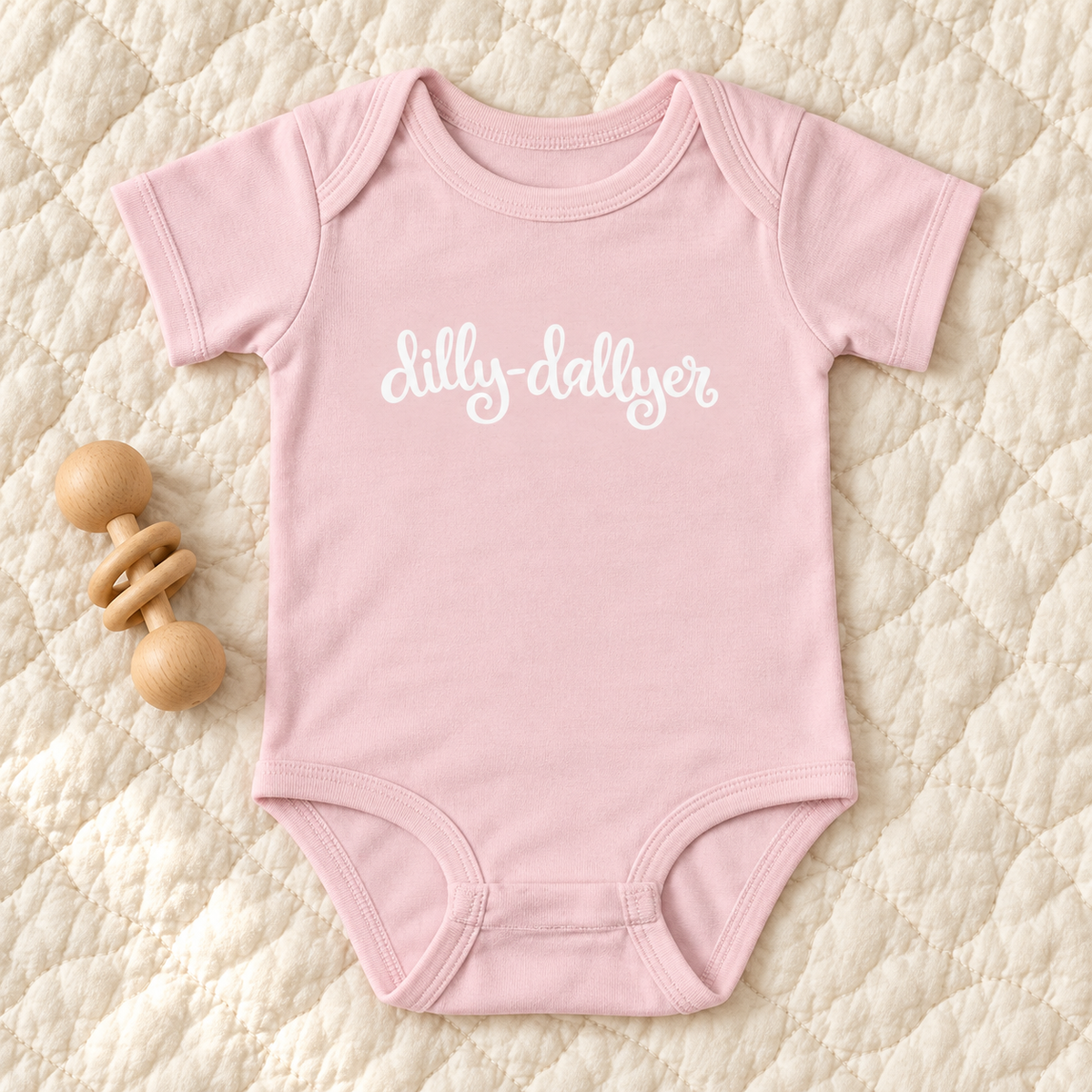 Ballerina pink baby bodysuit with white “dilly-dallyer” script beside a wooden rattle on a cream quilt