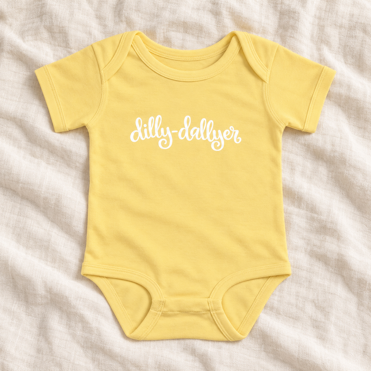 Banana baby bodysuit with white “dilly-dallyer” script, laid flat on ivory linen