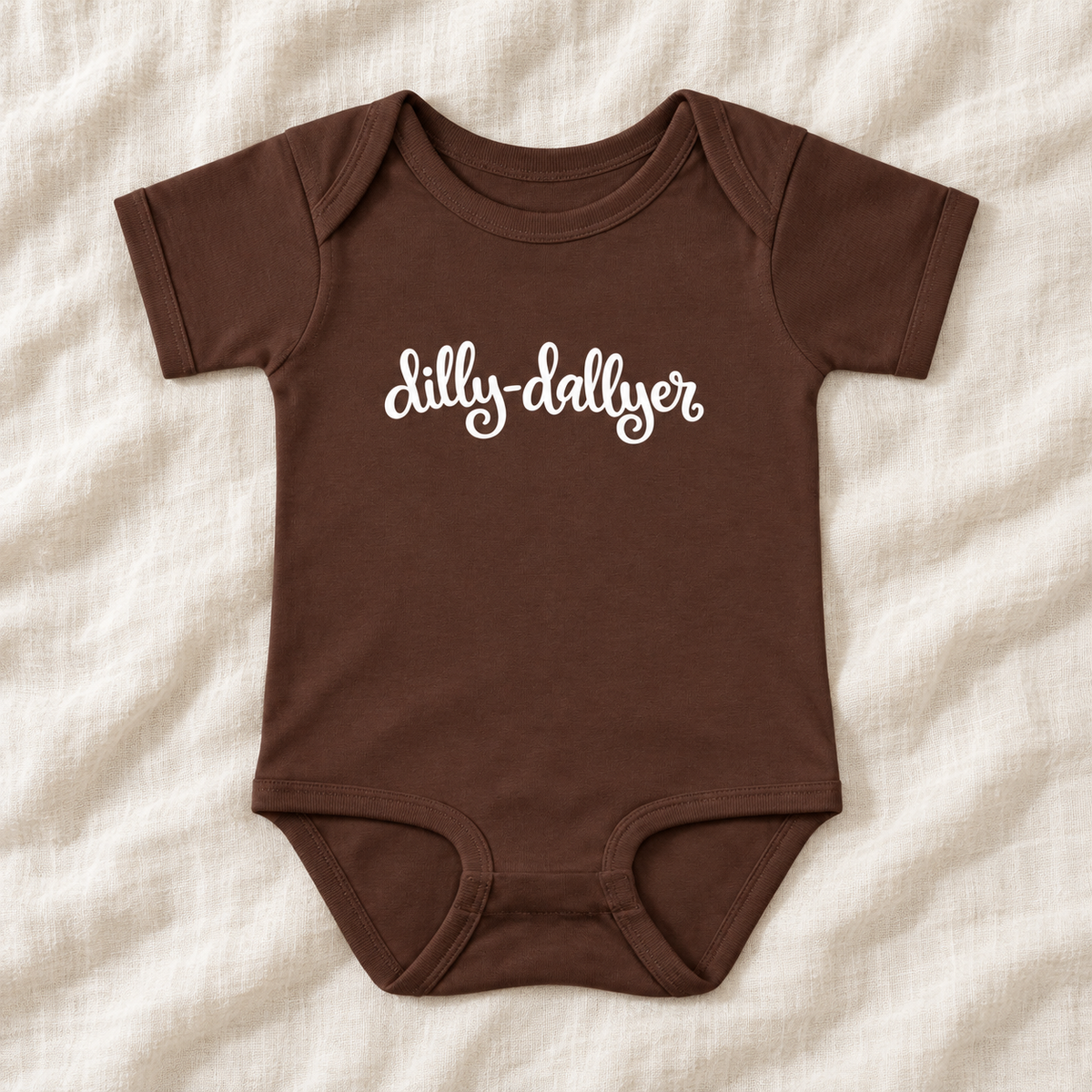 Brown baby bodysuit with white “dilly-dallyer” script, laid flat on ivory linen