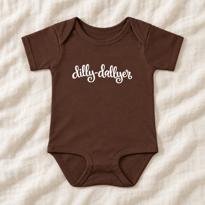 Brown baby bodysuit with white “dilly-dallyer” script, laid flat on ivory linen