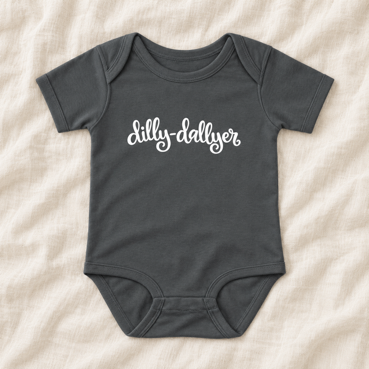Charcoal baby bodysuit with white “dilly-dallyer” script, laid flat on ivory linen