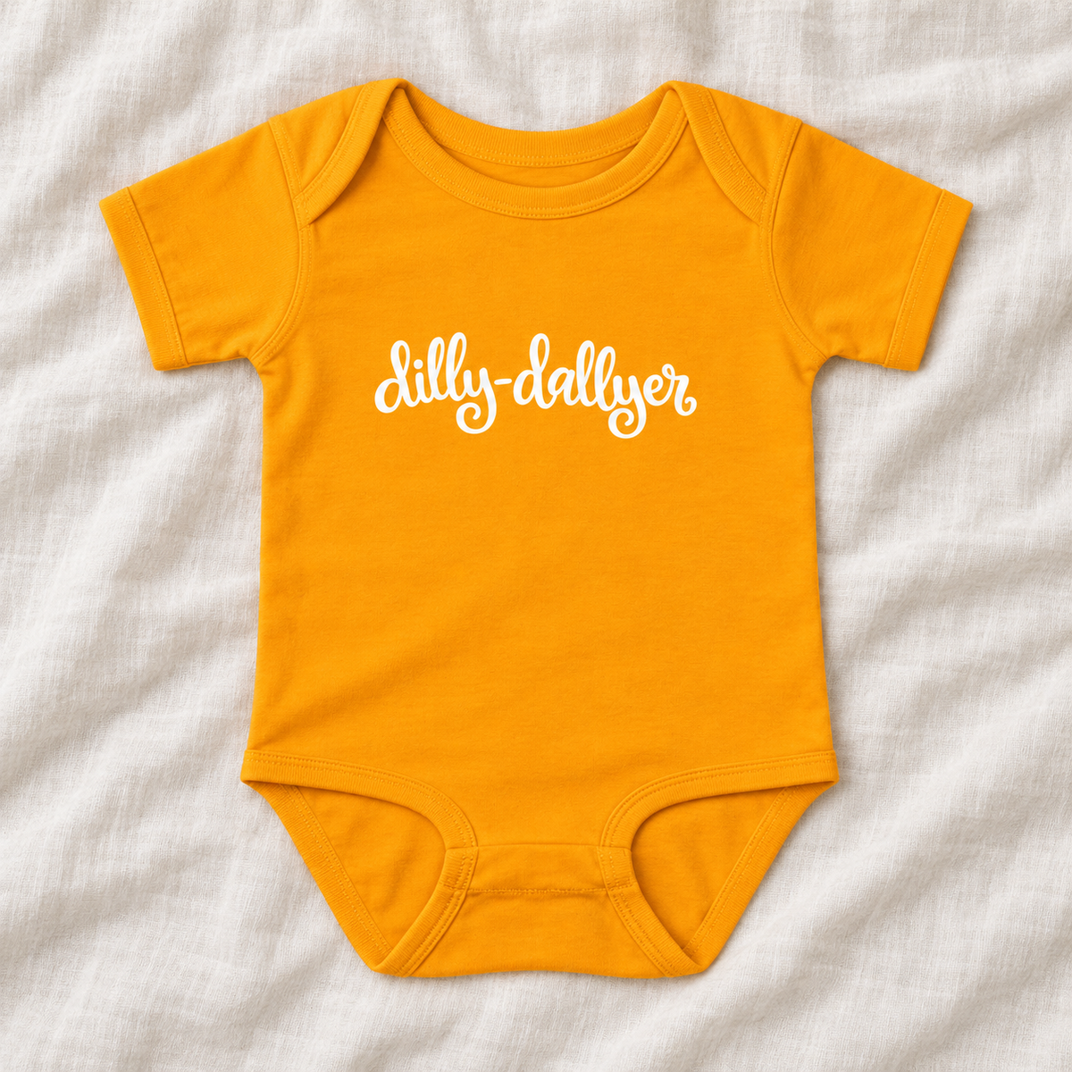 Gold baby bodysuit with white “dilly-dallyer” script, laid flat on ivory linen