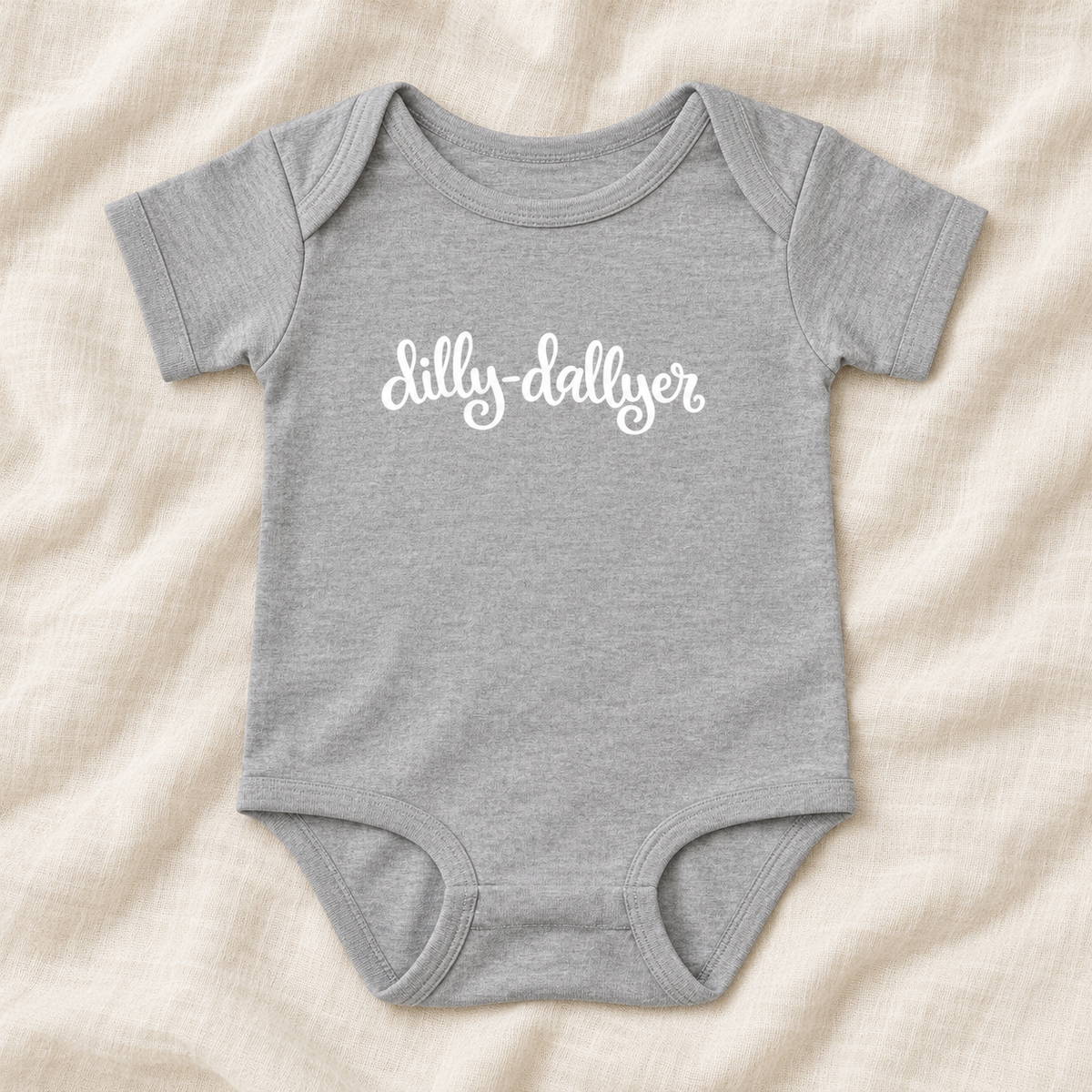 Heather baby bodysuit with white “dilly-dallyer” script, laid flat on ivory linen