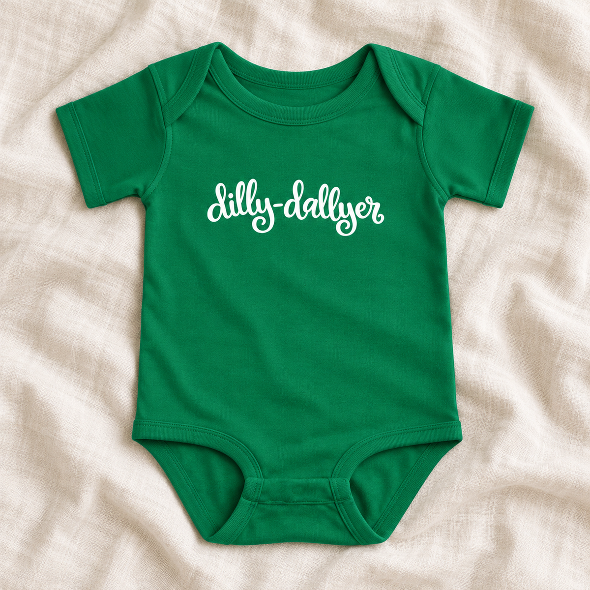 Kelly baby bodysuit with white “dilly-dallyer” script, laid flat on ivory linen