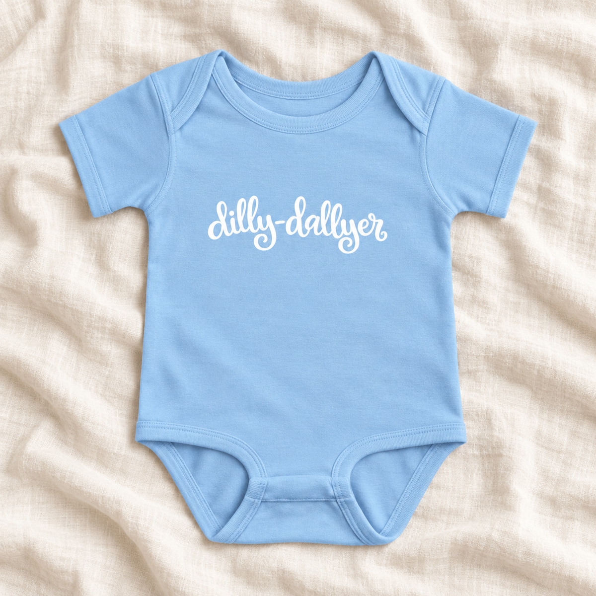 Light Blue baby bodysuit with white “dilly-dallyer” script, laid flat on ivory linen