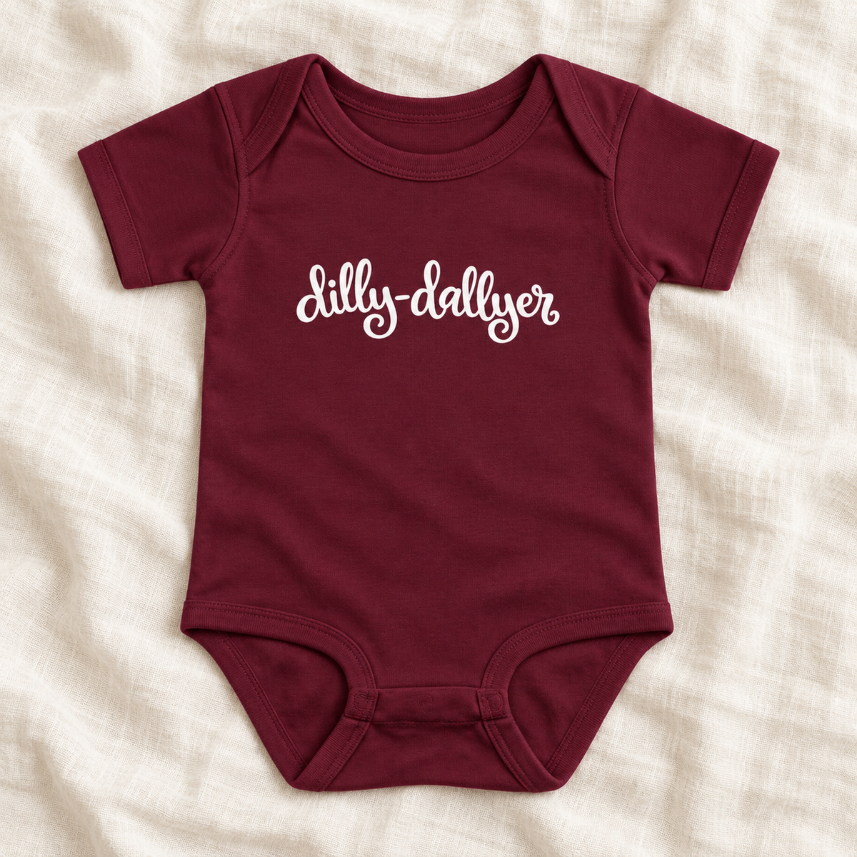 Maroon baby bodysuit with white “dilly-dallyer” script, laid flat on ivory linen
