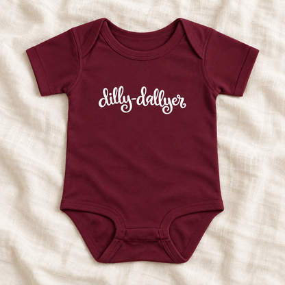 Maroon baby bodysuit with white “dilly-dallyer” script, laid flat on ivory linen