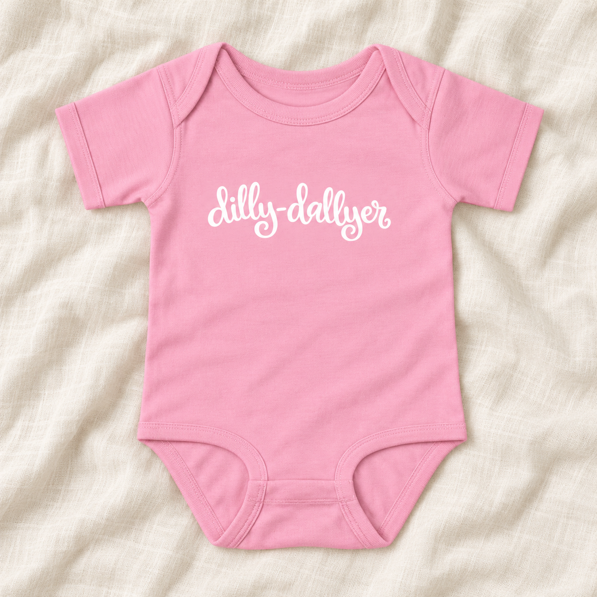 Pink baby bodysuit with white “dilly-dallyer” script, laid flat on ivory linen