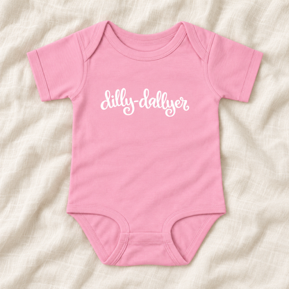 Pink baby bodysuit with white “dilly-dallyer” script, laid flat on ivory linen