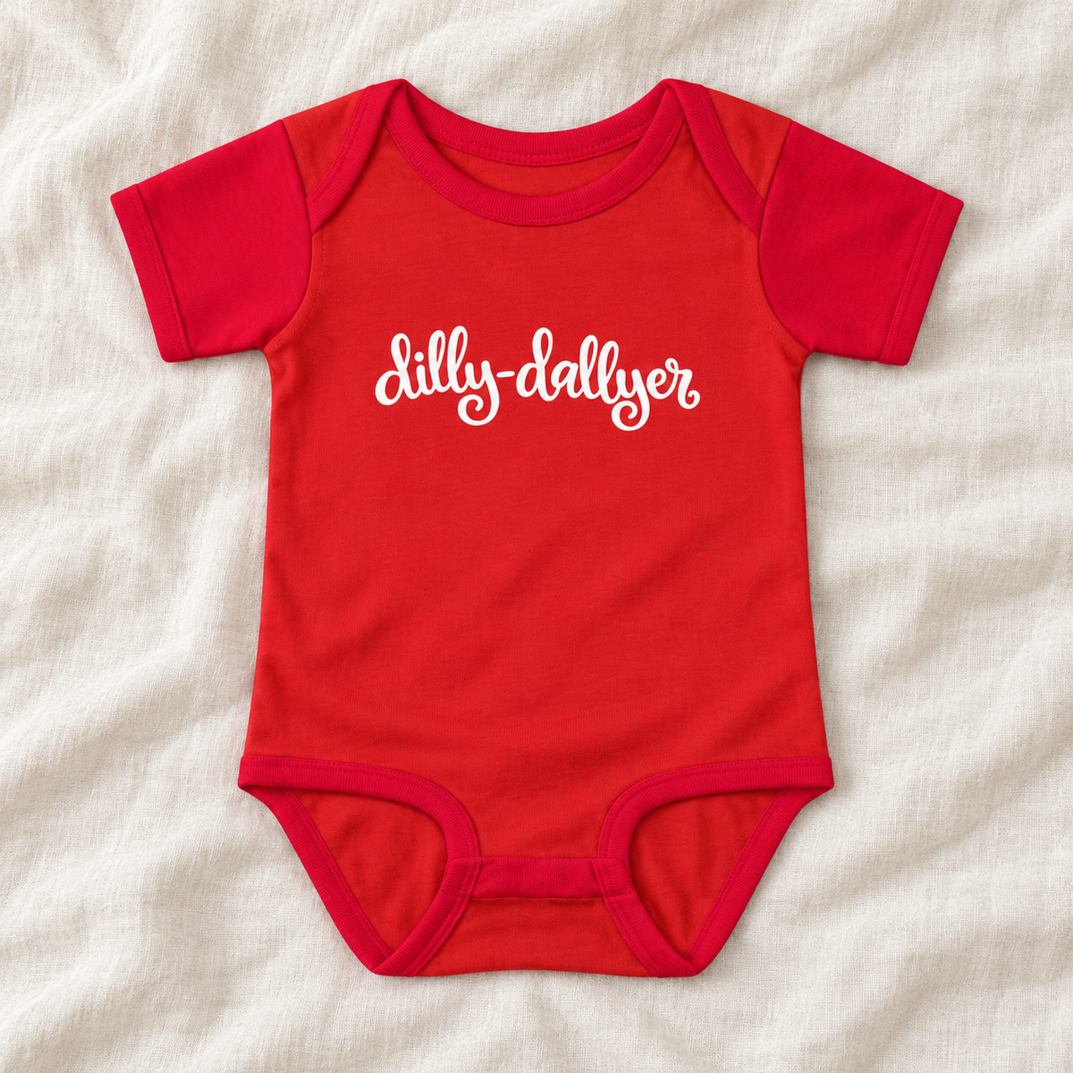 Red baby bodysuit with white “dilly-dallyer” script, laid flat on ivory linen
