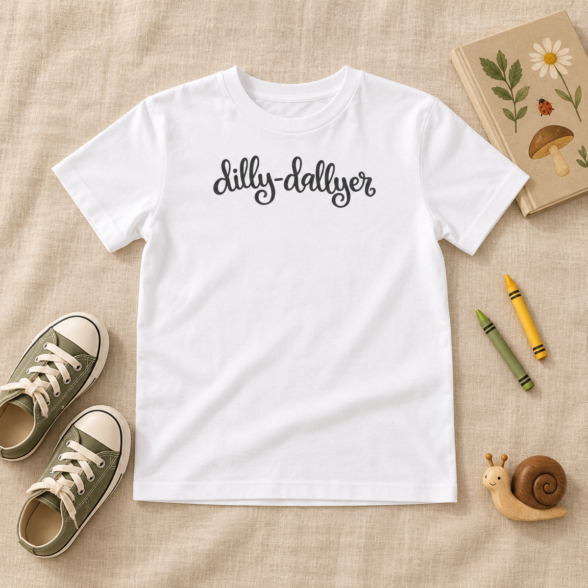 White Dilly-Dallyer kids tee beside sneakers, crayons, a nature book, and toy snail