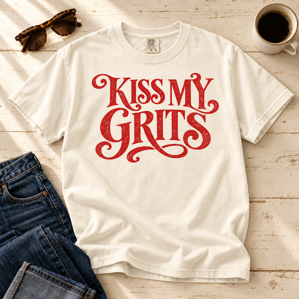 Ivory Kiss My Grits cotton tee laid flat with jeans, sunglasses, and coffee.