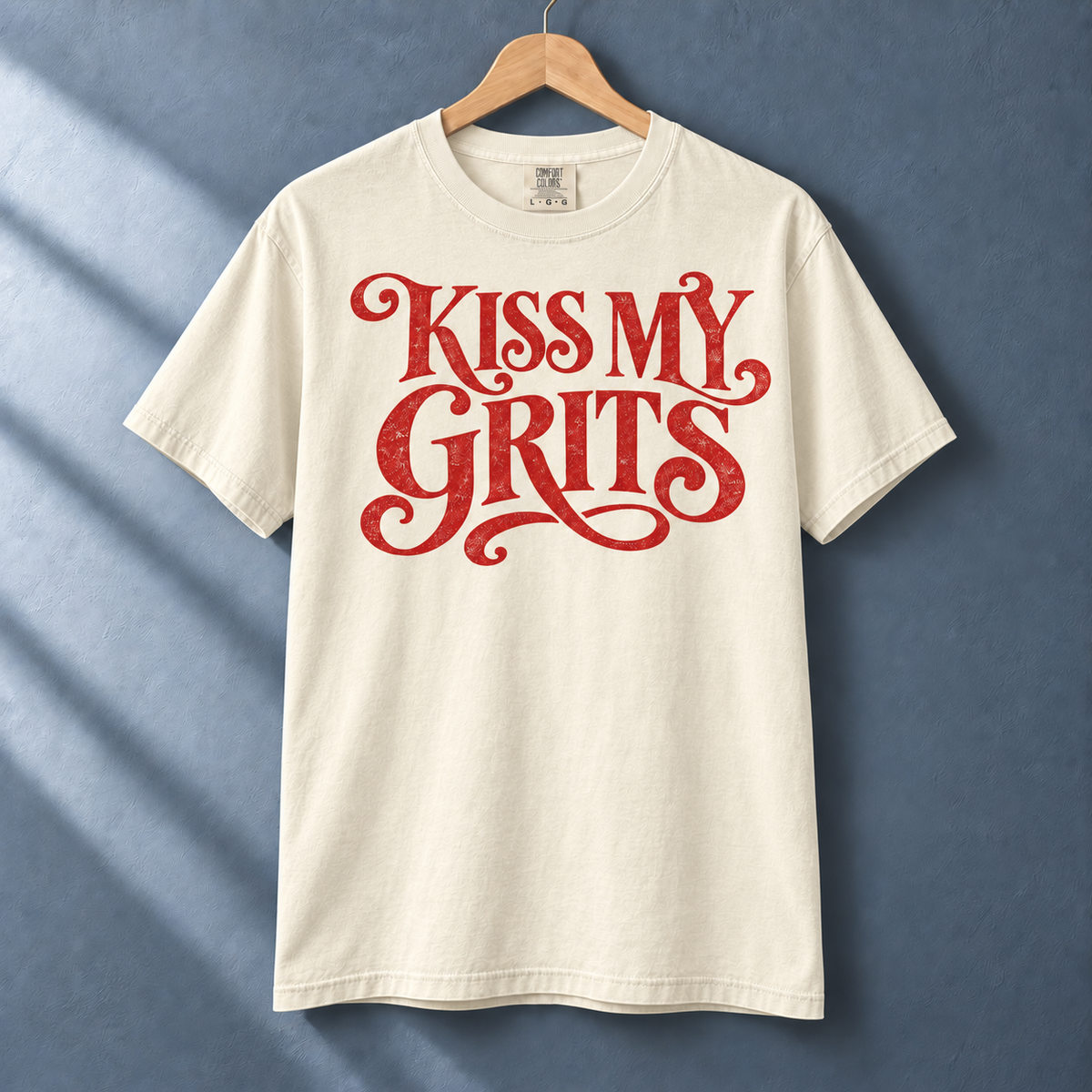 Ivory Kiss My Grits graphic tee with red vintage lettering on a hanger against a blue-gray wall.