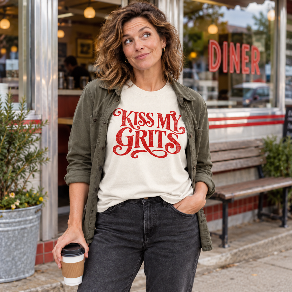 Woman wearing an ivory Kiss My Grits tee and olive jacket outside a diner.
