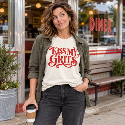 Woman wearing an ivory Kiss My Grits tee and olive jacket outside a diner.