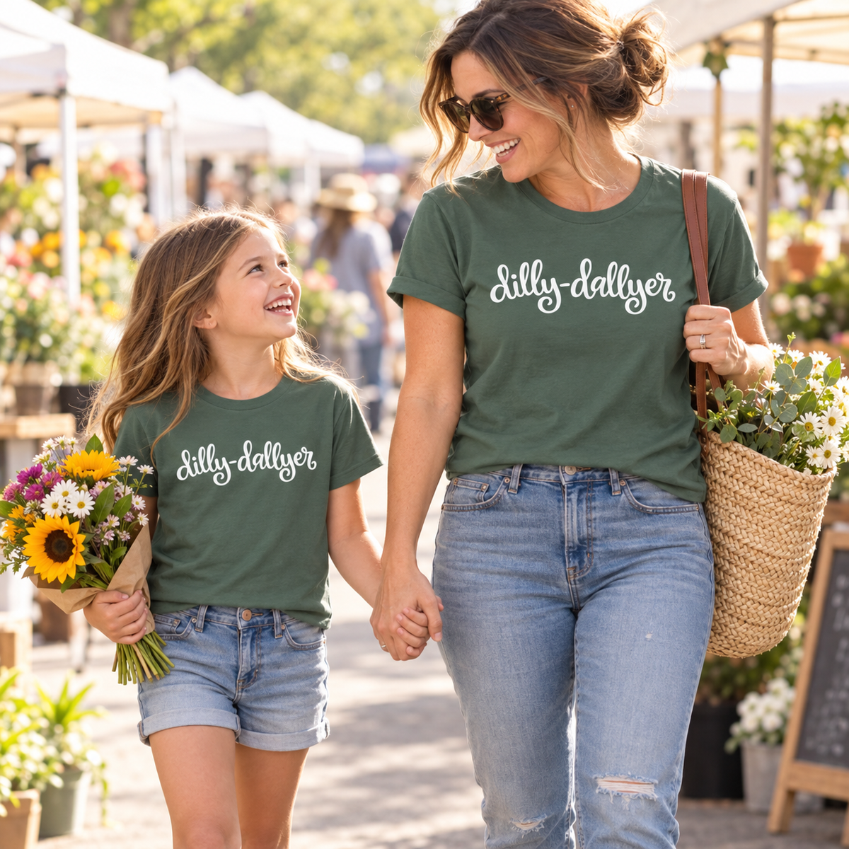 Mother and daughter wearing matching green "dilly-dallyer" T-shirts at a sunny farmers market.