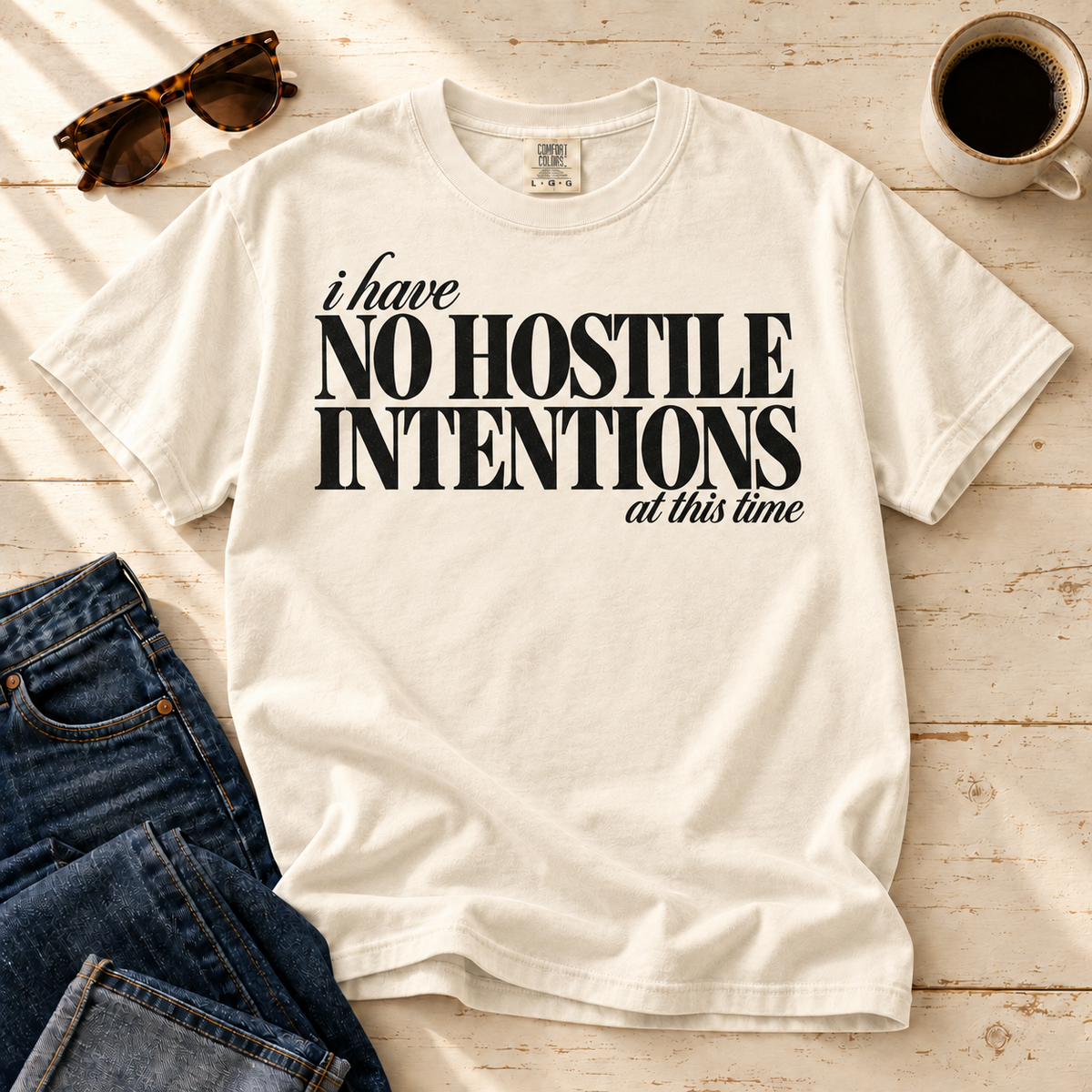 Ivory I Have No Hostile Intentions tee laid flat with jeans, sunglasses, and coffee.