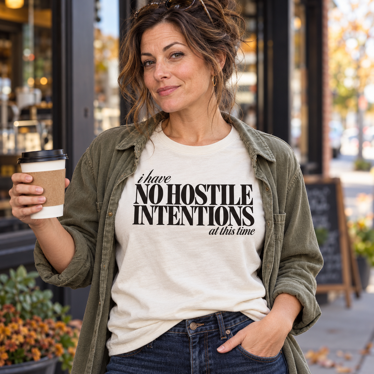 Woman in an ivory I Have No Hostile Intentions tee and olive overshirt holding coffee outdoors.