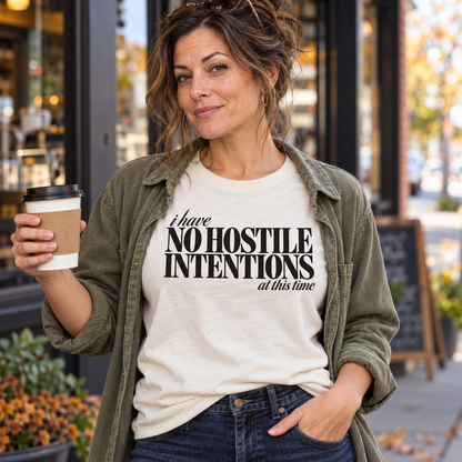 Woman in an ivory I Have No Hostile Intentions tee and olive overshirt holding coffee outdoors.