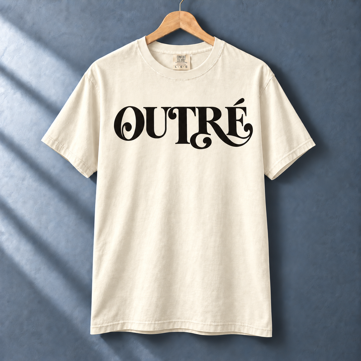 Ivory Outré tee with black flourished typography on a hanger against a blue-gray wall.