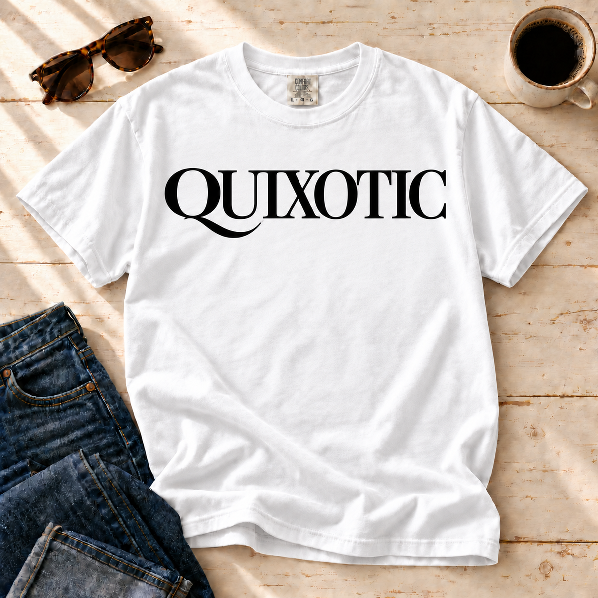 White Quixotic tee laid flat with jeans, sunglasses, and coffee.
