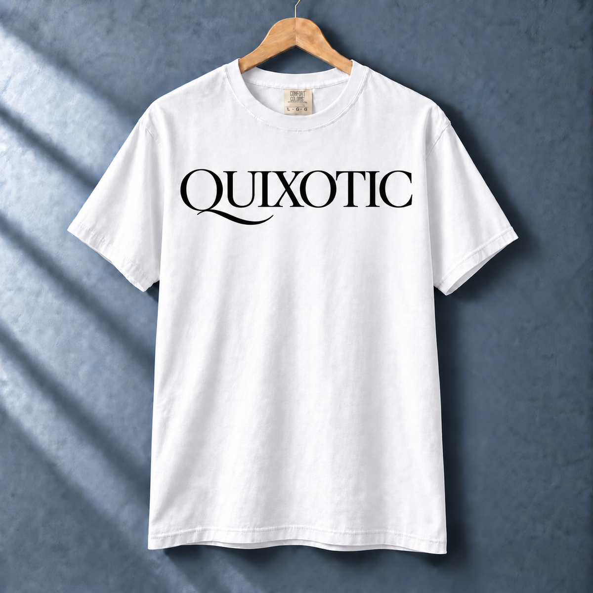 White Quixotic tee with black serif lettering and Q flourish on a hanger against a blue-gray wall.